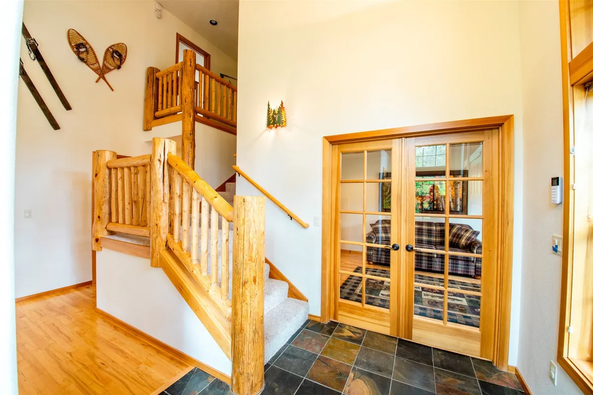 An interior scene highlighting a cozy living area with a plaid couch, wooden staircase, glass double doors, and decorative skis and snowshoes. The decor immediately connects to outdoor recreation, and the double doors imply direct access from inside to the adventure waiting outside.