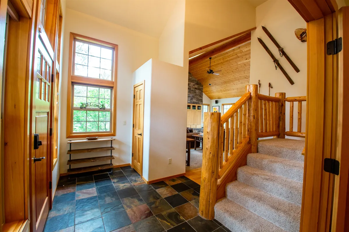 This image captures the home's inviting entryway, featuring a wooden staircase, slate flooring, large windows, and a substantial wooden door. Natural light streams in, creating a warm atmosphere and offering a glimpse of the surrounding resort environment.