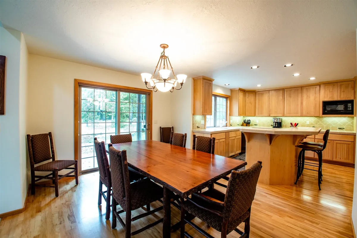 The spacious dining area features a large wooden table, a prominent chandelier, expansive windows with plenty of sunlight, and adjacent kitchen cabinetry. The setup encourages group gatherings, productive meetings, or casual meals after a day of work and recreation.