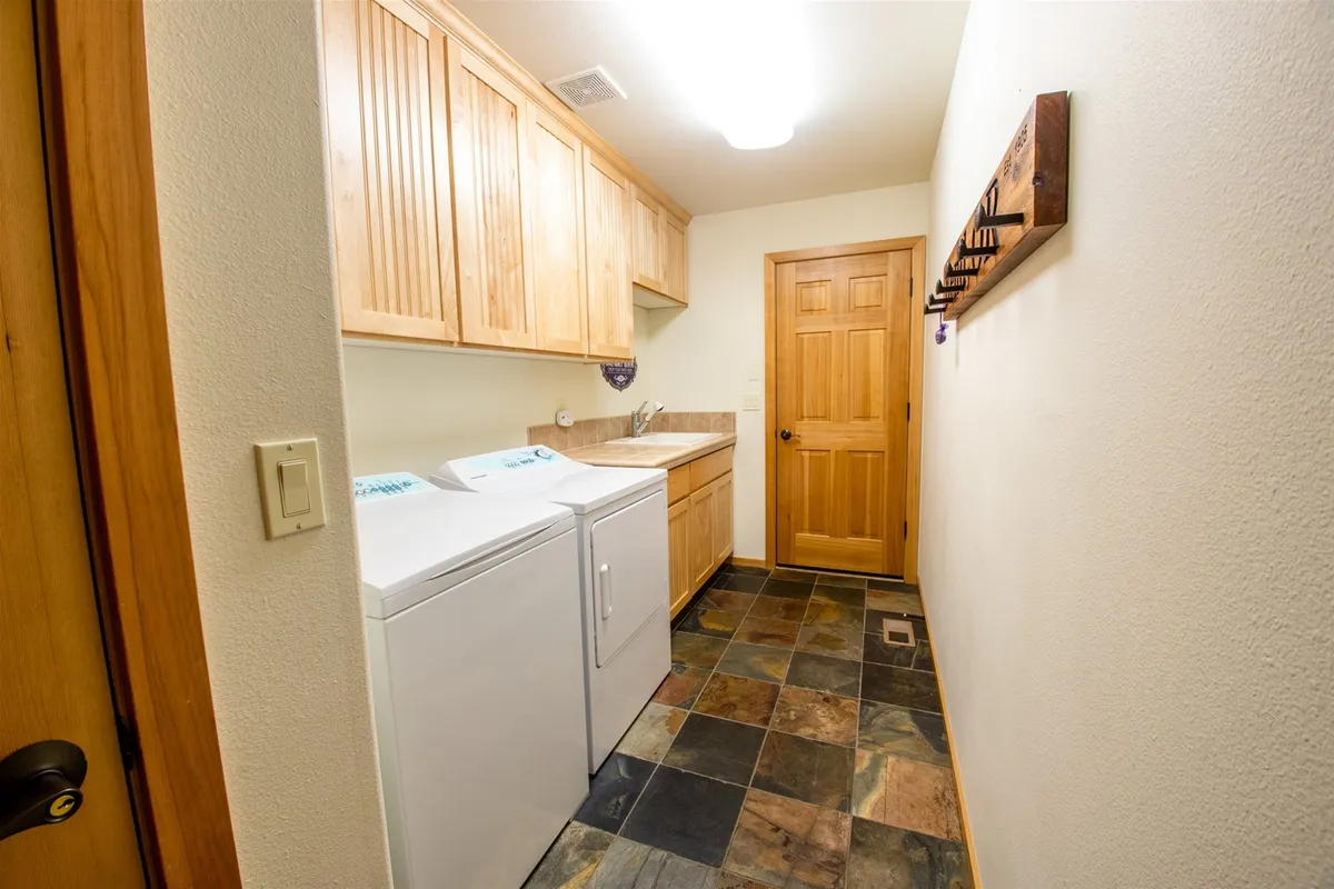 A laundry room featuring a modern washing machine and dryer, wooden cabinetry for storage, a coat rack, and a door leading to another room. The space is organized, clean, and well-lit, emphasizing convenience for everyday tasks or longer stays.