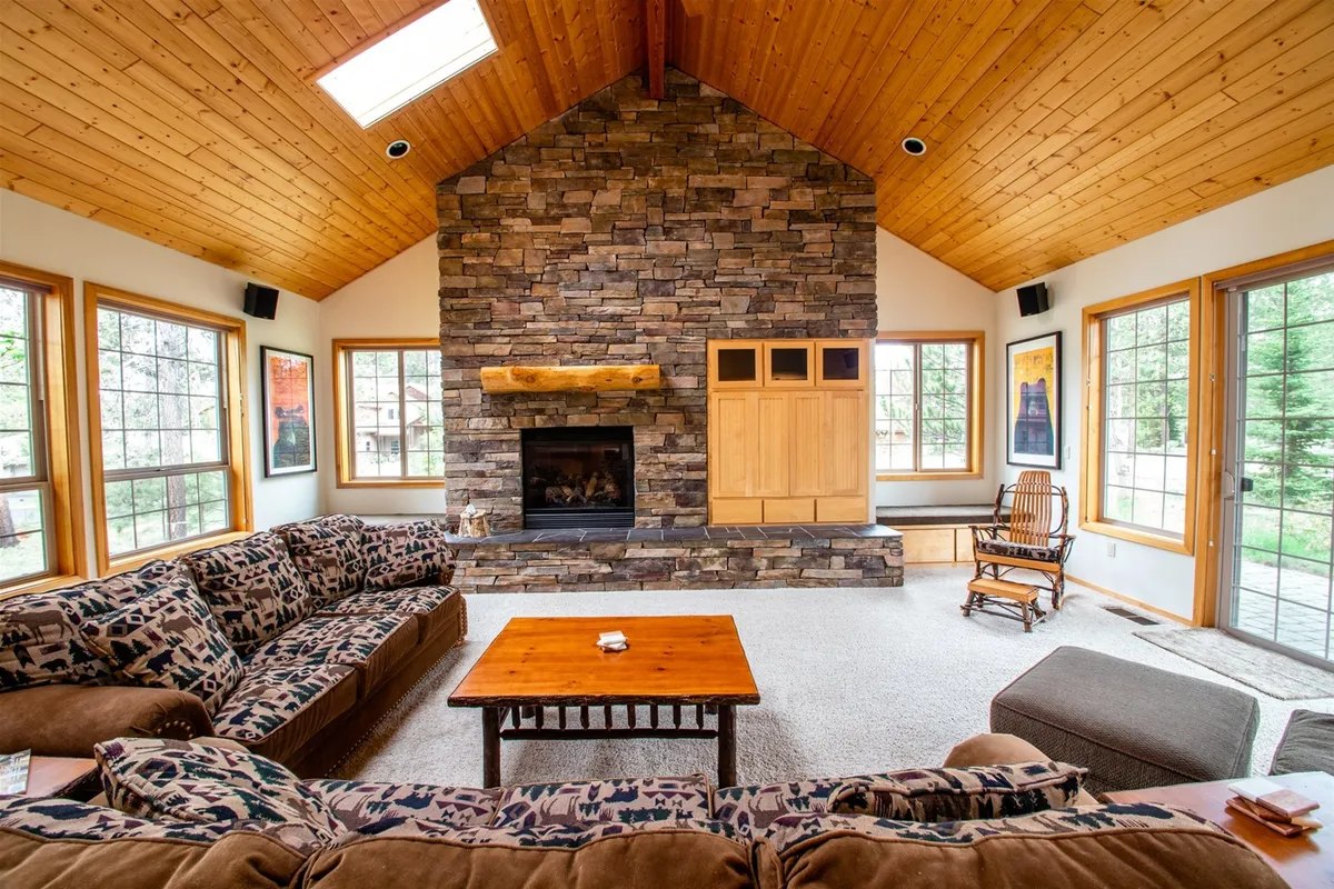 The image showcases a cozy living room with a striking stone fireplace as the focal point, surrounded by comfortable seating and bathed in natural light from large windows. A rustic wooden coffee table anchors the space, creating a warm, inviting atmosphere perfect for gatherings.