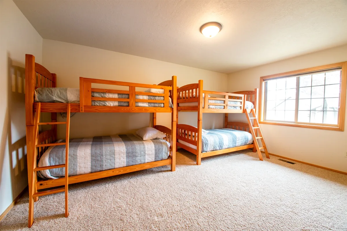 A cheerful bunk room featuring multiple bunk beds, a large window, and carpeted flooring. The room exudes a playful yet comfortable atmosphere, ideal for children’s sleepovers, bedtime stories, and playtime.