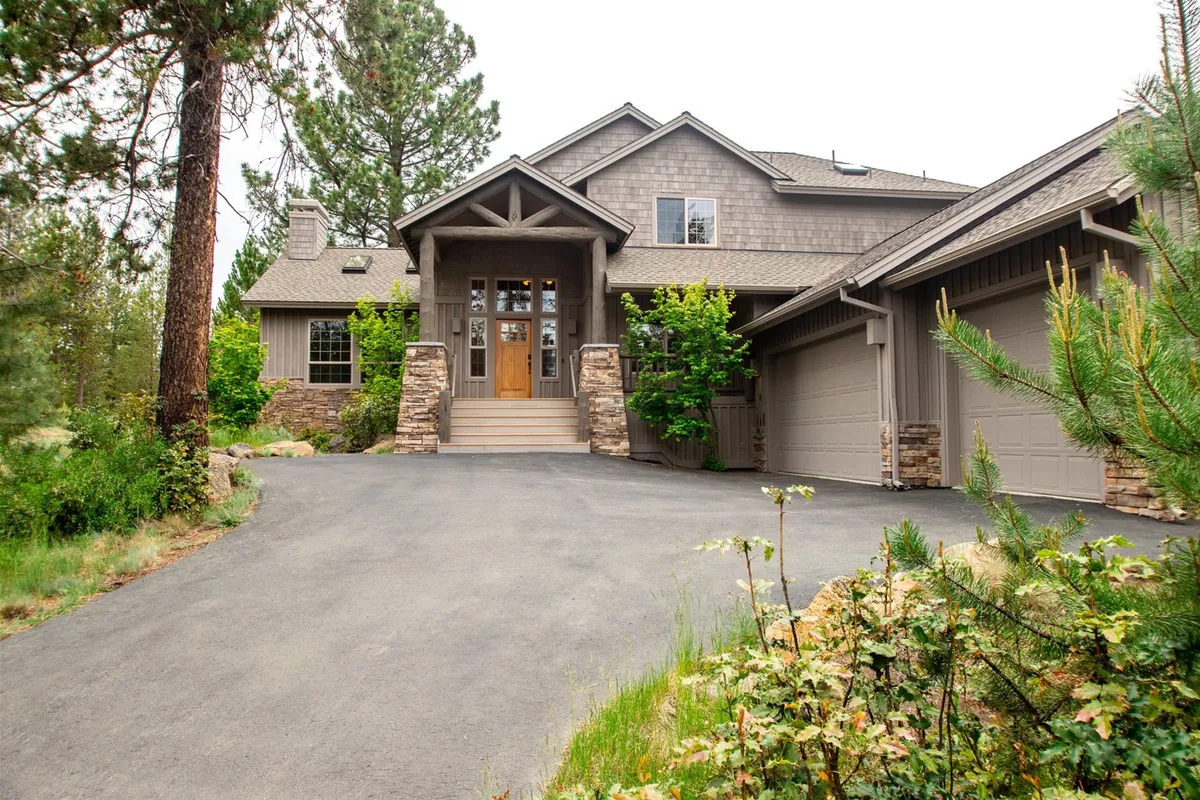 The image captures the front entrance of Tokatee 14, featuring stone pillars, a landscaped garden, and a welcoming porch. The outdoor setting is framed by greenery, creating an inviting first impression that hints at the home's proximity to Sunriver's outdoor adventures.