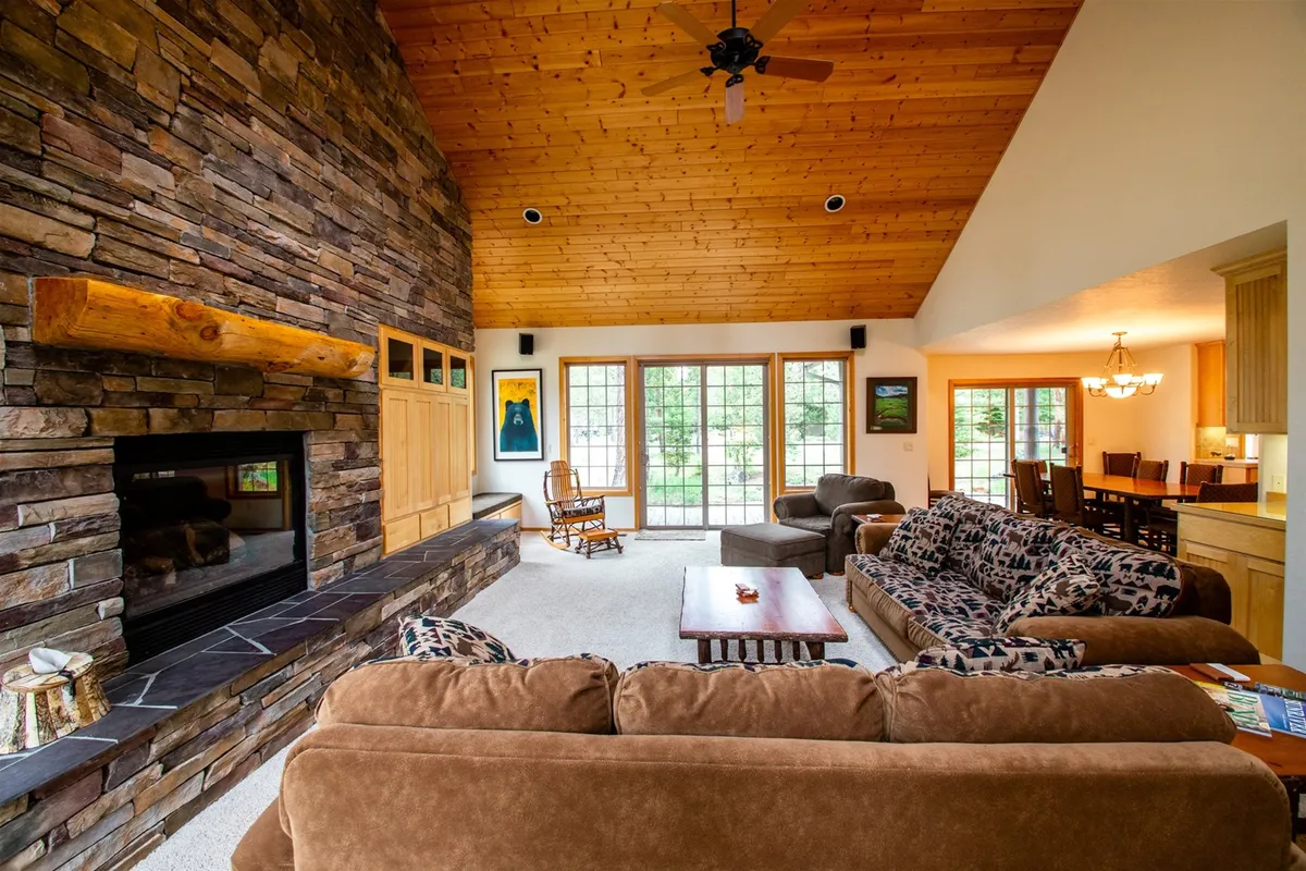 This image highlights a spacious open-concept living area featuring a striking stone fireplace, large windows letting in ample sunlight, plush sofas, and a visible dining area. The wood ceiling and natural tones evoke warmth and artistry, while the arrangement encourages gatherings and relaxation.
