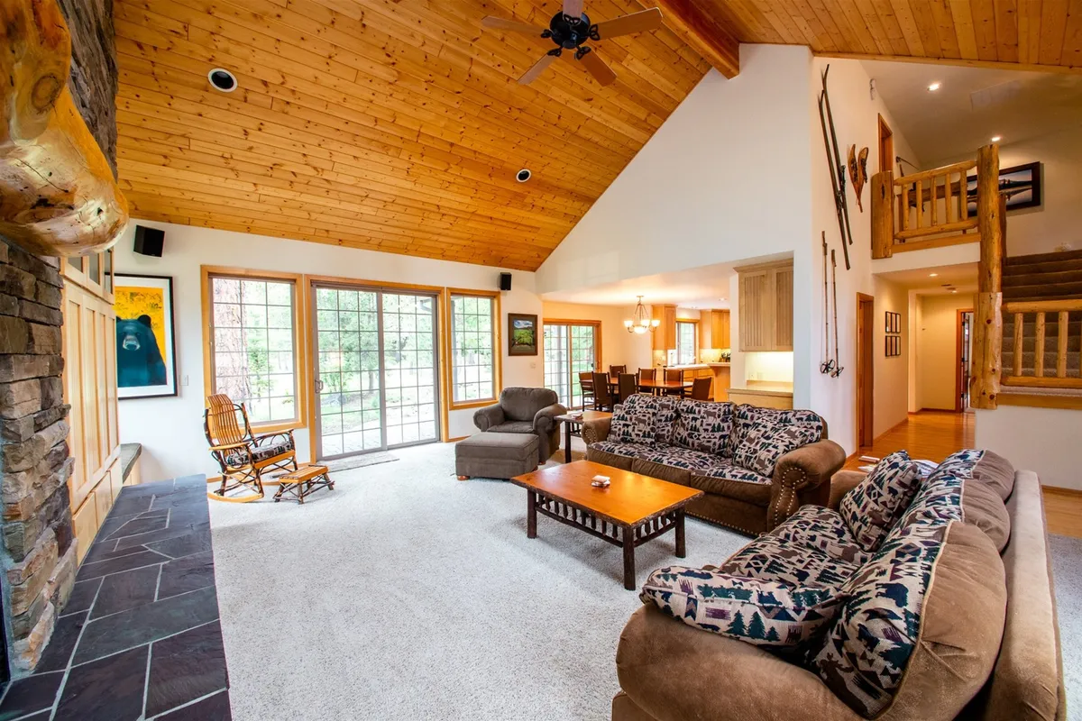 A wide-angle view of the open living space, showing the stone-faced fireplace, expansive windows, a cozy seating area, and a large dining table. The natural light floods the room, and there's a seamless connection to the outdoors, suggesting proximity to patio doors.