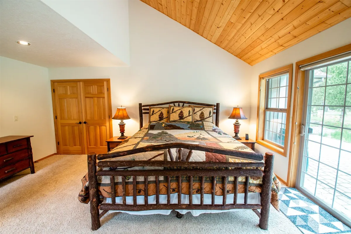A tranquil bedroom featuring a rustic wooden bed with a colorful quilt, warm bedside lighting, and a large window streaming in natural light. The decor is cozy and inviting, capturing the comfort of a premium sleeping space.