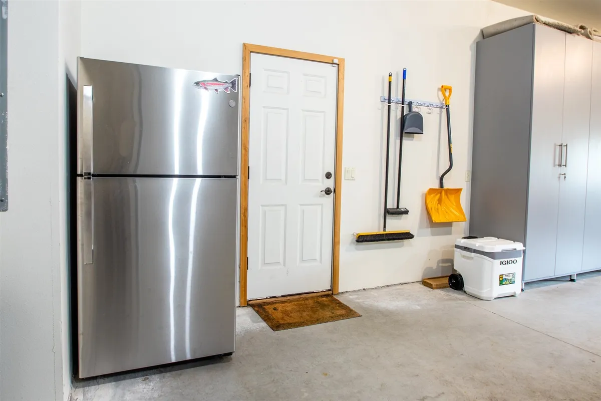 A tidy and well-organized garage space with a stainless steel refrigerator, white door, storage cabinets, and a clean concrete floor. The setting suggests easy storage and preparation for various recreational activities, hinting at gear and equipment ready for use.