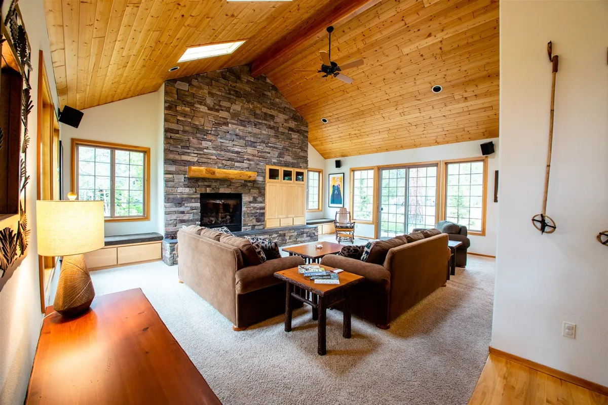 A warmly lit living area with a striking stone fireplace, large windows letting in natural light, wooden ceiling, and comfortable seating. The space is arranged for relaxation, social gatherings, and cozy evenings spent together.